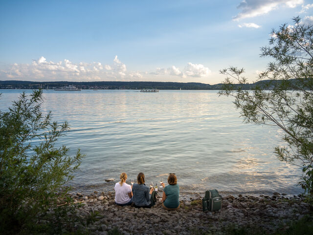 Drei Personen mit Weingläsern sitzen am Ufer des Bodensees mit Blick auf das Wasser. Im Hintergrund sind Bäume und zwei Schiffe sichtbar.